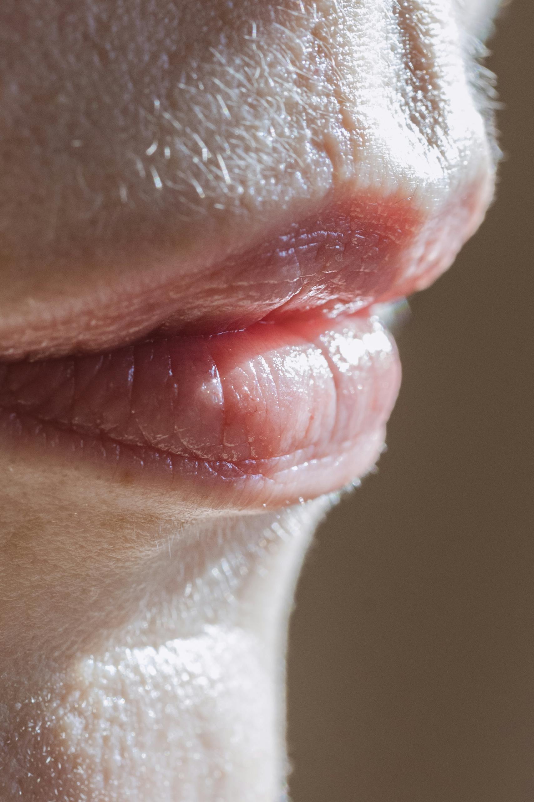 Closeup pink lips of crop unrecognizable female with short face hair standing in light room with bright sunlight on brown background