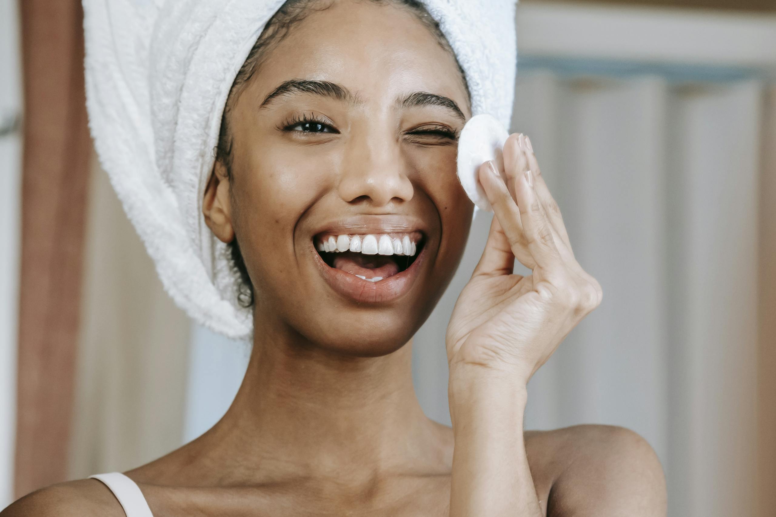 Delighted ethnic woman laughing brightly while looking at reflection and cleaning face with cotton pad in morning