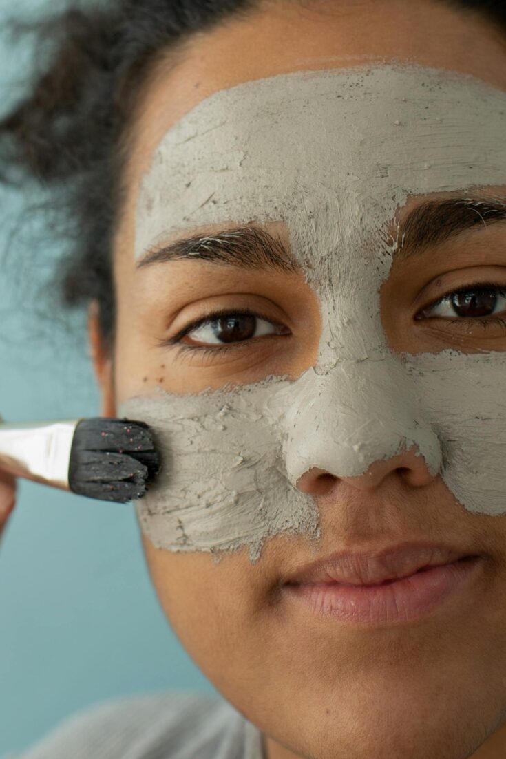 Content crop African American female applying clay facial mask with brush for skincare routine while looking at camera on blue background