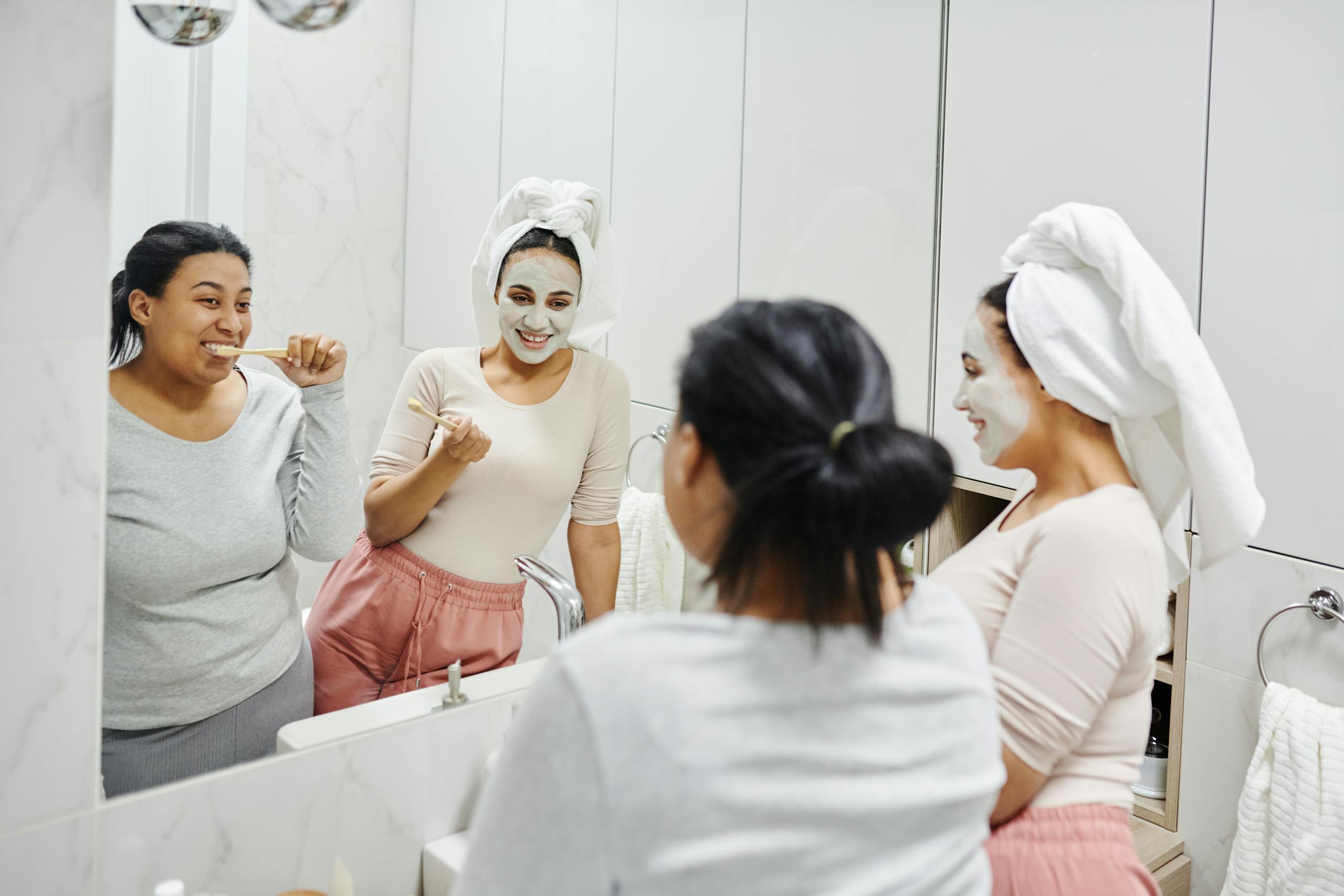 Woman With Head Towel Wearing Clay Mask Standing Beside Woman Brushing Her Teeth